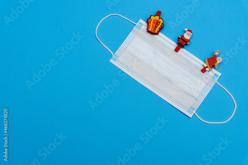 Medical mask on blue background in the upper right corner with pins - toys in the form of gift, Santa Claus and angel. Top view with copy space, space for text. Happy New Year 2022 concept.