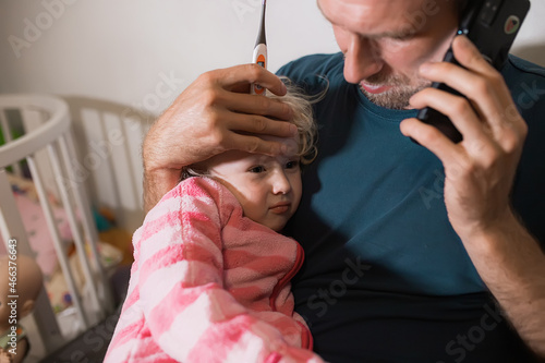 Dad keeps his hand on his daughter's forehead with fever during illness and helps to heal by calling the doctor and informing him about the illness or the flu by phone