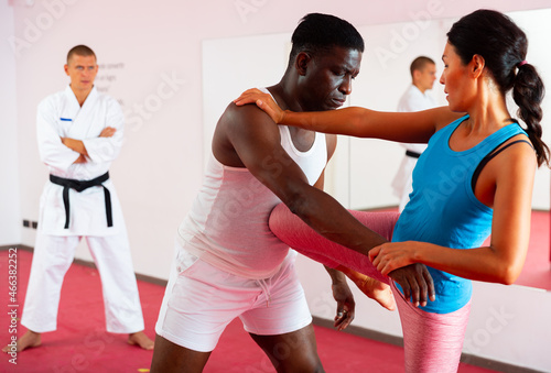 Asian woman exercising fighting moves with African-american man during self-defence training, Caucasian man in kimono standing and watching.