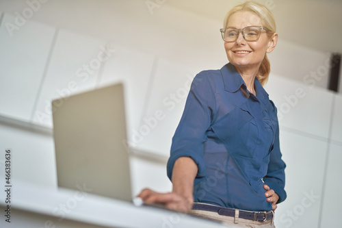 Portrait of successful mature businesswoman in glasses smiling, working on the laptop, standing in modern kitchen at home photo