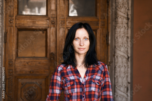 Close-up face of pretty woman with blue eyes, natural dark hair, no makeup, standing in a plaid shirt smiling, looking at the camera. Against the background of a building with wooden doors © Екатерина Переславце