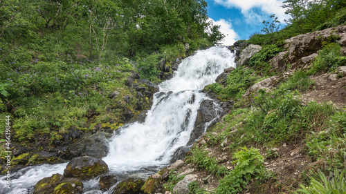 The stream flows down the mountain slope. The water is bubbling and foaming. On the banks - lush green grass  wildflowers. Boulders in the foreground. Blue sky. Kamchatka.