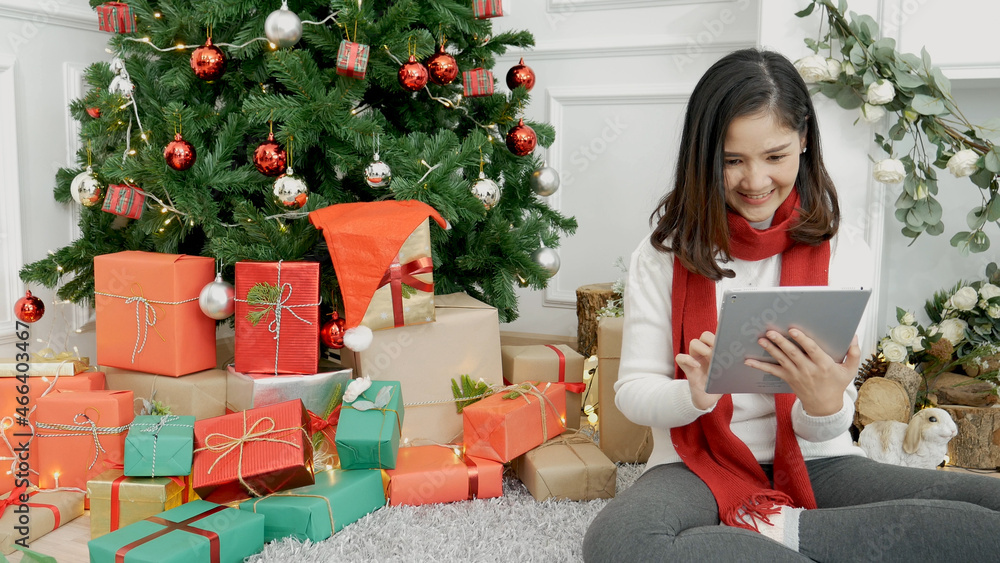 Asian woman using smart tablet sit at christmas tree xmas present gift box in cozy living room. Woman hand touch screen smile shopping online, happiness time christmas holiday winter season festive Asian woman using smart tablet sit at christmas tree xmas present gift box in cozy living room. Woman hand touch screen smile shopping online, happiness time christmas holiday winter season festive