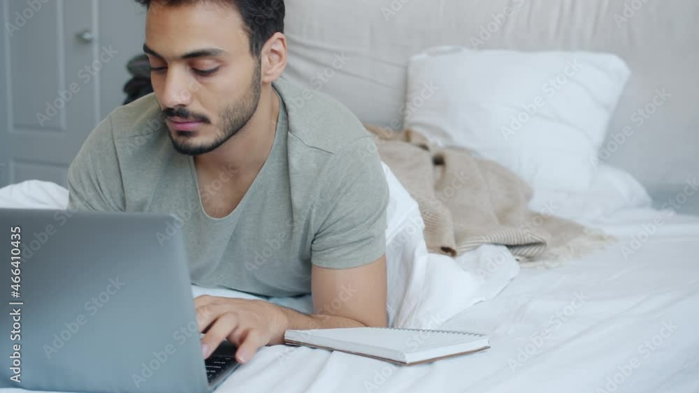 Middle Eastern man student is using laptop typing and writing in notebook lying in comfy bed alone. Online education and technology concept. Middle Eastern man student is using laptop typing and writing in notebook lying in comfy bed alone. Online education and technology concept.
