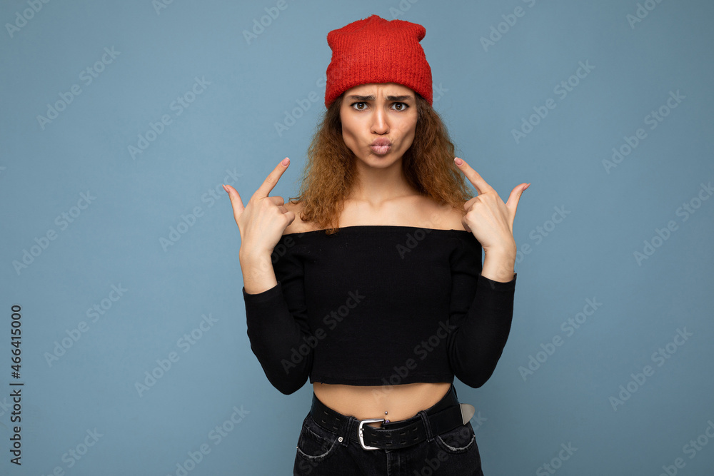 Photo of young positive beautiful brunette curly woman with sincere emotions wearing black crop top and red hat isolated on blue background with copy space and having fun Photo of young positive beautiful brunette curly woman with sincere emotions wearing black crop top and red hat isolated on blue background with copy space and having fun