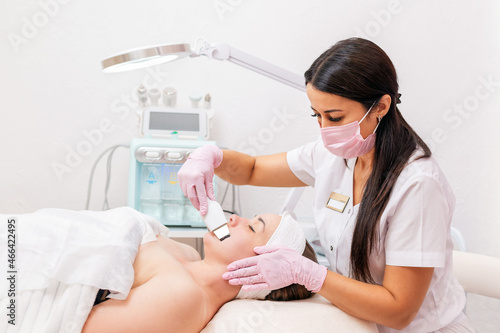 Beauty salon. A cosmetologist wearing a medical mask and rubber gloves cleans the face using an ultrasound machine. In the background is a cosmetic device. Concept of professional cosmetology
