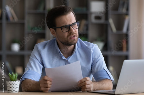 Stressed young businessman in eyewear feeling nervous of getting bad news in paper correspondence, reading letter with bank loan rejection or bankruptcy notification, having financial problems. photo