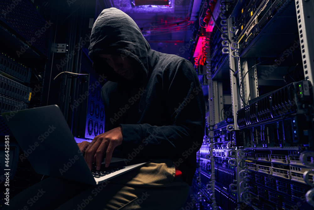 Hacker seated in server room launching cyberattack on laptop Hacker seated in server room launching cyberattack on laptop