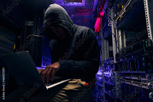 Hacker seated in server room launching cyberattack on laptop