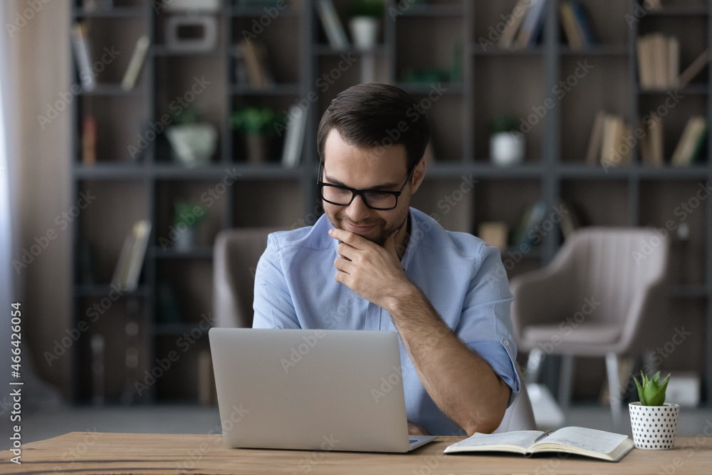 Smiling young businessman in eyewear looking at laptop screen, reading email with pleasant news, feeling satisfied with marketing research results, working on online project in modern office room. Smiling young businessman in eyewear looking at laptop screen, reading email with pleasant news, feeling satisfied with marketing research results, working on online project in modern office room.