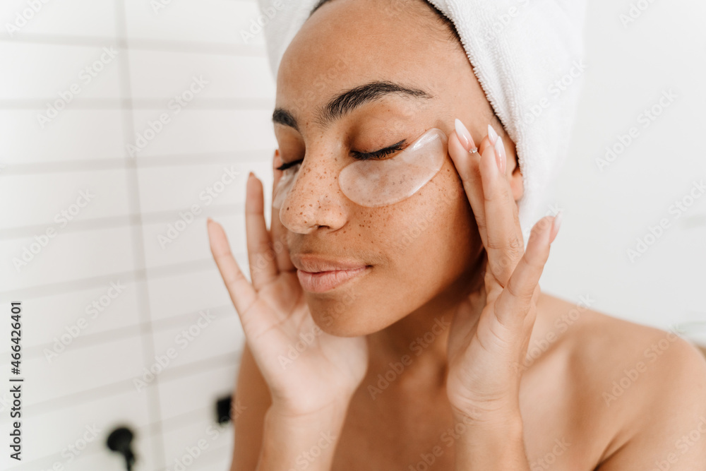 Young black woman wrapped in towels applying eye patches Young black woman wrapped in towels applying eye patches