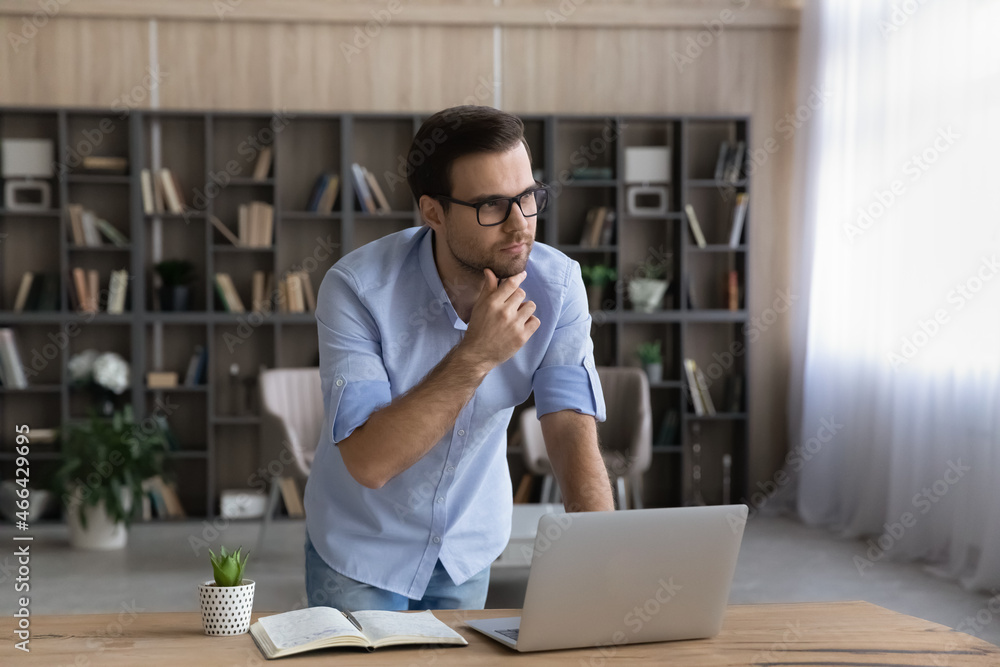 Thoughtful young businessman manager in eyeglasses looking in distance, considering problem solution, creating new online project on computer standing at table in modern home office workplace. Thoughtful young businessman manager in eyeglasses looking in distance, considering problem solution, creating new online project on computer standing at table in modern home office workplace.