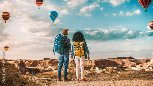 Young Diverse Tourist Couple Hiking with Backpacks in Great Wilderness in Rocky Canyon Valley. Male and Female Holding Hands on Adventure Trip. Hot Air Balloon Festival in Mountain National Park.