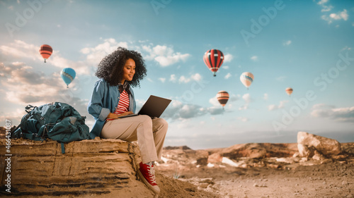 Obraz na plátně Excited Beautiful Young Multiethnic Female Traveller with Afro Hairstyle Using Laptop Computer on Top of Rocky Canyon Valley