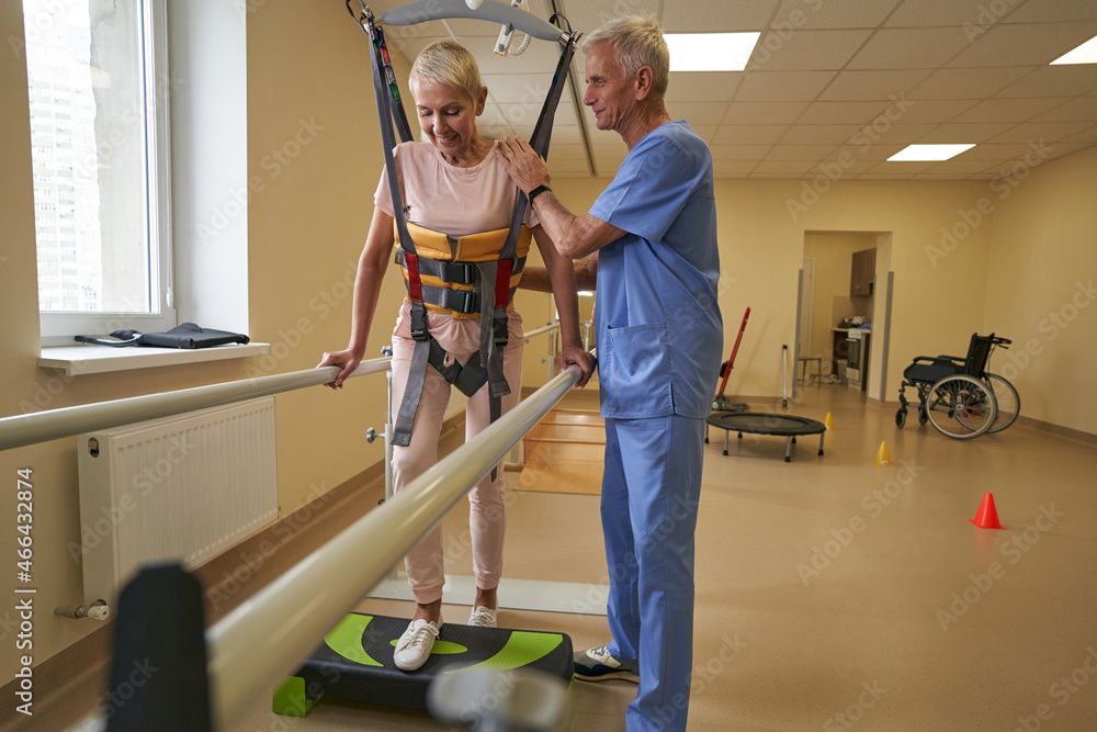 Mature woman walking between parallel bars at rehabilitation room