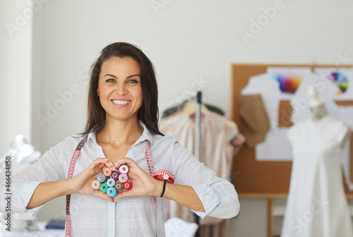 Portrait of friendly seamstress woman showing hands heart to camera in sewing workshop. Woman who does her favorite job makes heart out of multicolored threads and wears pins with measuring tape. photo