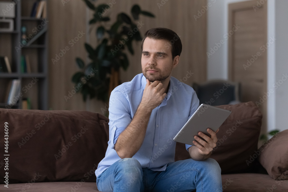 Thoughtful young handsome man looking in distance holding digital tablet in hands, web surfing information, spending free leisure time online, sitting on comfortable sofa in modern living room.