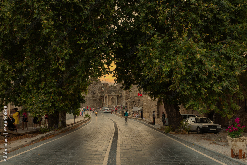 Fototapeta premium Views of the stone paved road leading from the entrance gate of the ancient city of Side towards the city center. Cloudy Sunset Sky and Sea view. Dust, Dark and Shadow