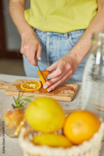Female hand cut orange, Woman preparing, making citrus and rosemary fresh lemonade in glass on a white table at home, summer drink, detox healthy water.