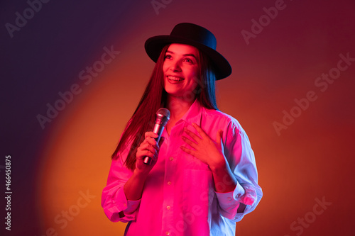One young beautiful girl wearing black hat using phone isolated on dark studio background in neon light. Concept of emotions, fashion
