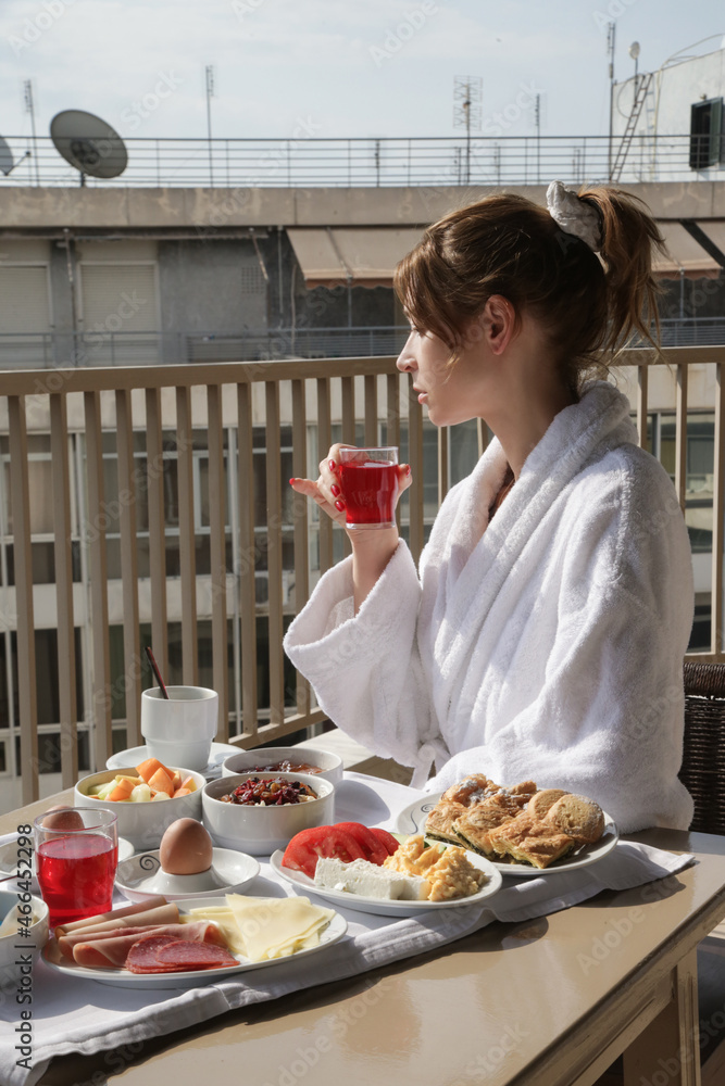 Young woman in bathrobe enjoying coffee and breakfast at the balcony with city view. Calm idyllic morning before busy day.