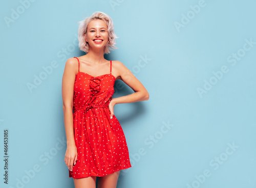 Portrait of young beautiful smiling female in trendy summer red dress. Sexy carefree woman posing near blue wall in studio. Positive blond model having fun and going crazy. Cheerful and happy