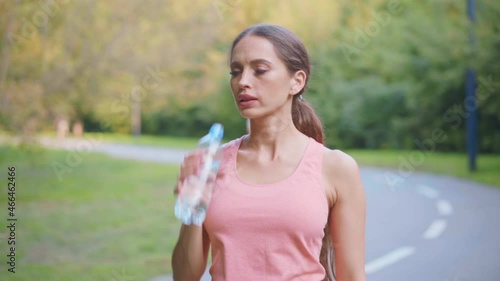 Female runner drink water transparent plastic bottle after morning jogging Caucasian athletic female resting after workout summer day in park medium shoot beautiful sportswoman quenches thirst photo