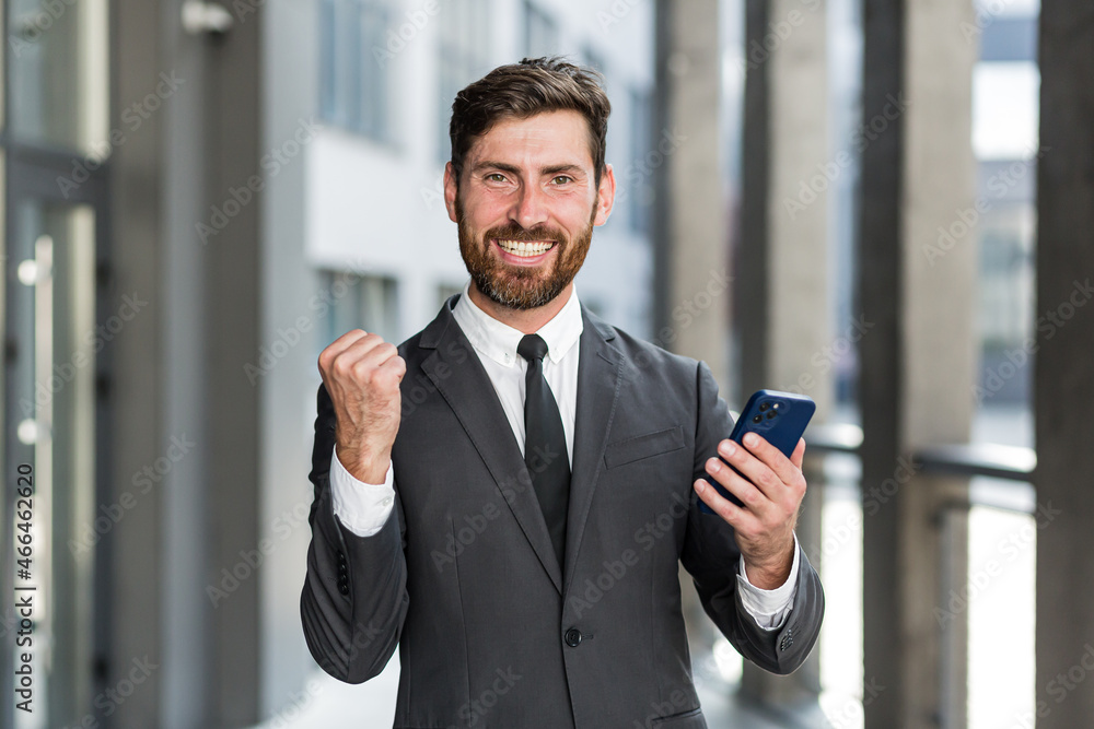 Successful and happy businessman shouts with joy and satisfaction of winning, reads good news from a mobile phone, outside office looks at camera Successful and happy businessman shouts with joy and satisfaction of winning, reads good news from a mobile phone, outside office looks at camera