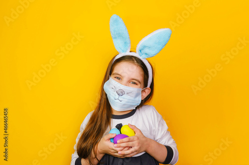 Girl kid 6 y.o. in rabbit bunny ears on head and protective mask with the face of an animal with colored eggs isolated on yellow studio background. Covid easter child. © NATALYA