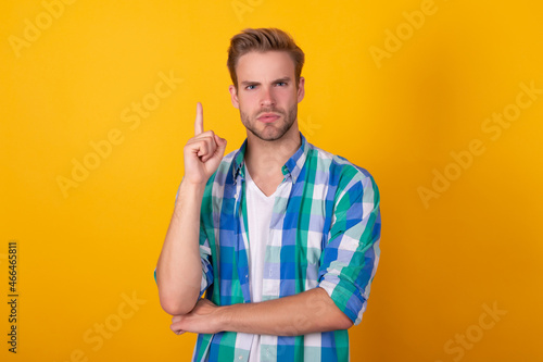 Serious handsome man in checked shirt keep raised finger pointing up yellow background, idea