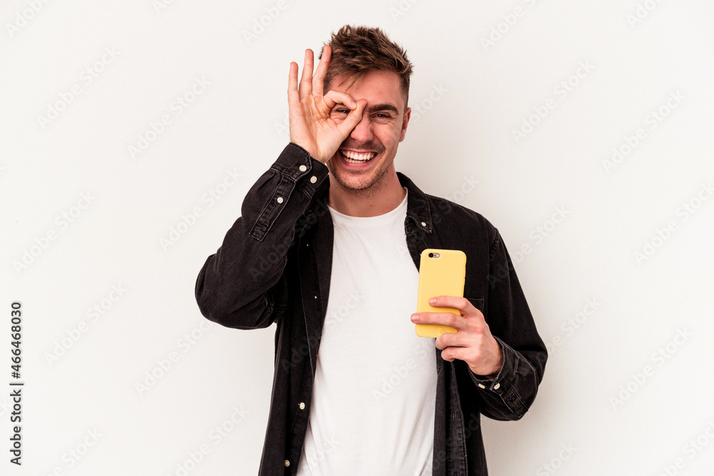 Young caucasian man holding a mobile phone isolated on white background excited keeping ok gesture on eye.