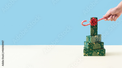 Trying to steal a gift from the top of a Christmas tree with candy cane. Green, red and gold decorations on yellow and blue pastel background. Christmas, winter, new year concept. 