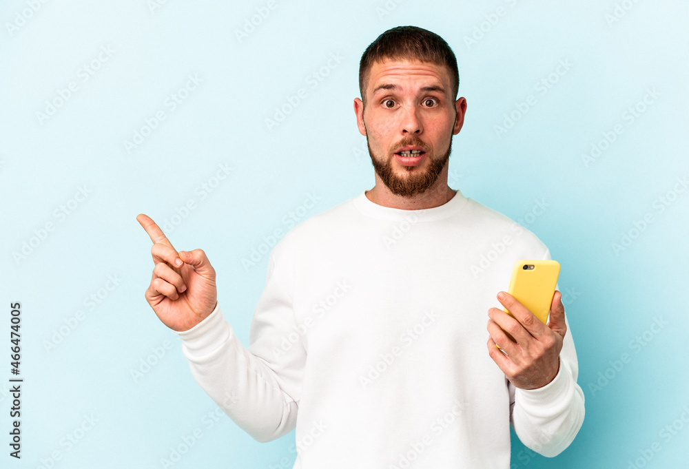 Young caucasian man holding mobile phone isolated on blue background pointing to the side Young caucasian man holding mobile phone isolated on blue background pointing to the side