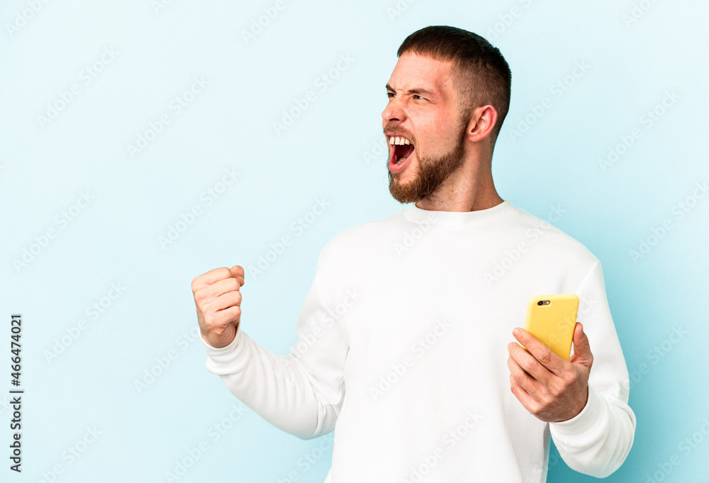 Young caucasian man holding mobile phone isolated on blue background raising fist after a victory, winner concept. Young caucasian man holding mobile phone isolated on blue background raising fist after a victory, winner concept.