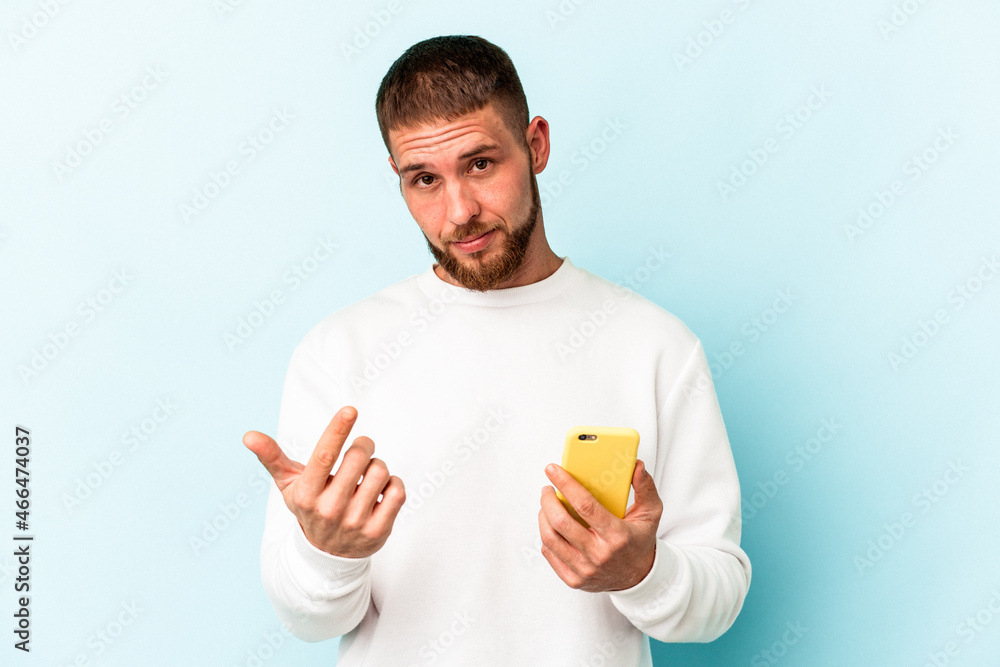 Young caucasian man holding mobile phone isolated on blue background pointing with finger at you as if inviting come closer. Young caucasian man holding mobile phone isolated on blue background pointing with finger at you as if inviting come closer.