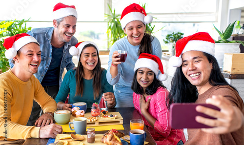 merry christmas! happy multiethnic group of friends wearing xmas santa hat making a selfie portrait sitting on a table at sweet breakfast time enjoying brunch together on winter festivities indoors