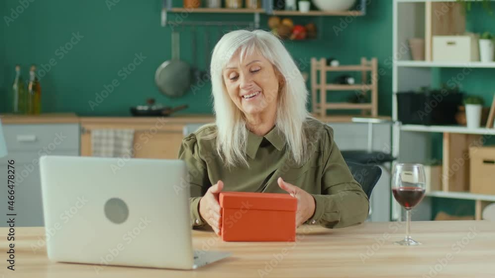 Happy Elderly Woman Celebrating Online Enjoying Online Conversation, Woman Enthusiastically Opening Gift Box While Chatting in the Living Room At Home. Positive Emotions and Gift Concept. Happy Elderly Woman Celebrating Online Enjoying Online Conversation, Woman Enthusiastically Opening Gift Box While Chatting in the Living Room At Home. Positive Emotions and Gift Concept.