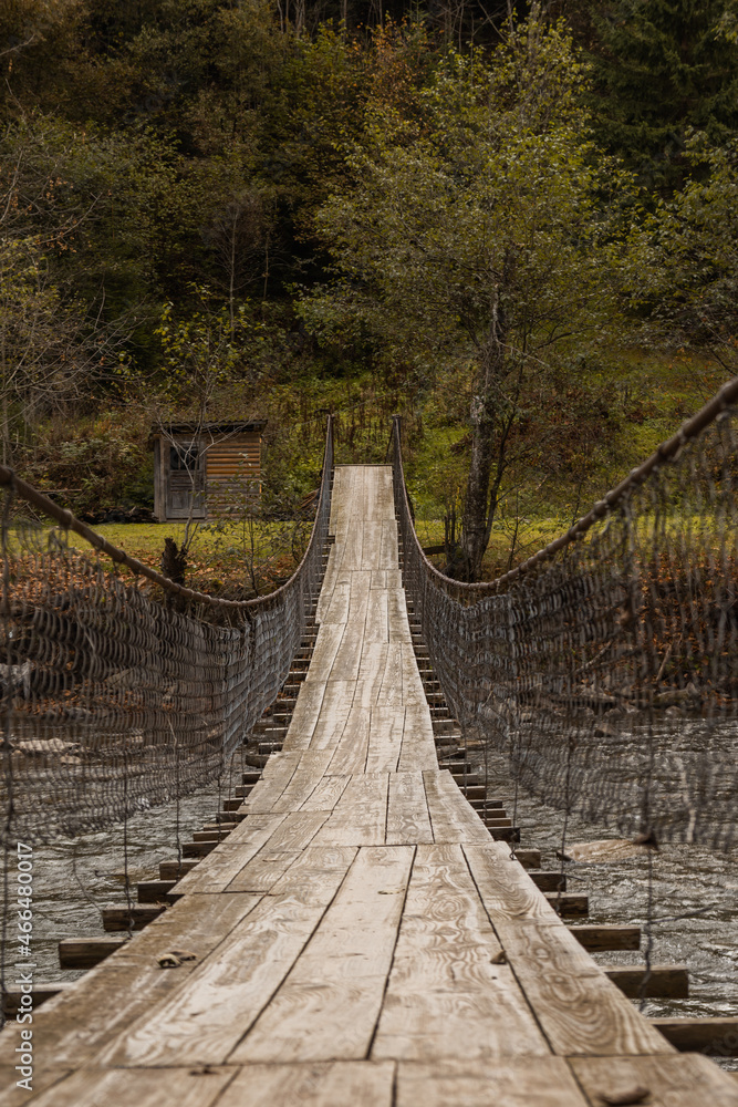 Obraz premium Old wooden suspension bridge across mountain river among autumn forest. Moody image. Countryside background.