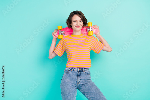 Photo of pretty funky young woman dressed striped t-shirt smiling holding skateboard isolated teal color background © deagreez