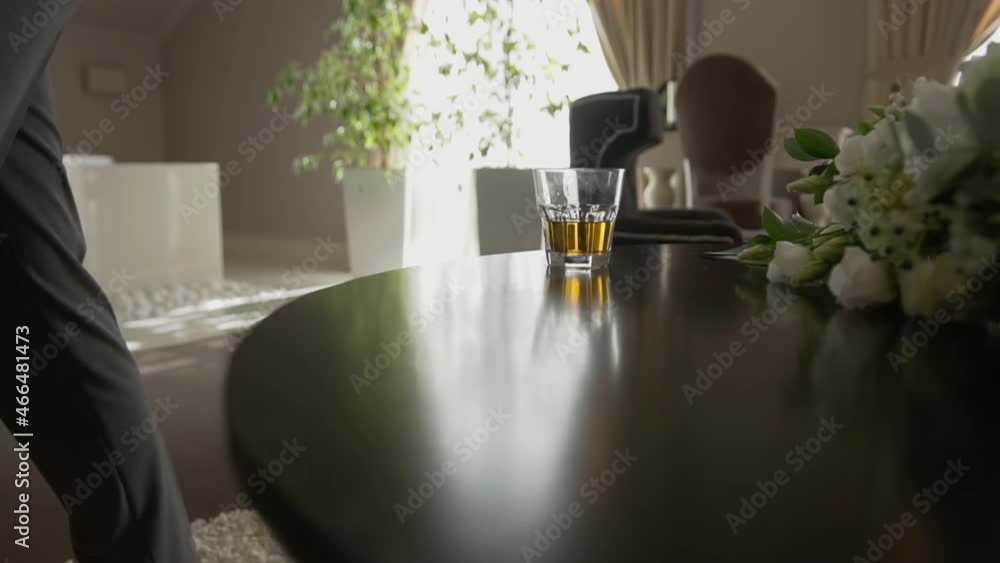Successful Caucasian man approaches wooden table and takes clean glass with expensive luxury drink. Two glasses with brown alcohol on table with flowers. Man starts important day with whiskey Successful Caucasian man approaches wooden table and takes clean glass with expensive luxury drink. Two glasses with brown alcohol on table with flowers. Man starts important day with whiskey