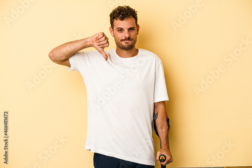 Young caucasian man holding crutch isolated on yellow background showing a dislike gesture, thumbs down. Disagreement concept. photo