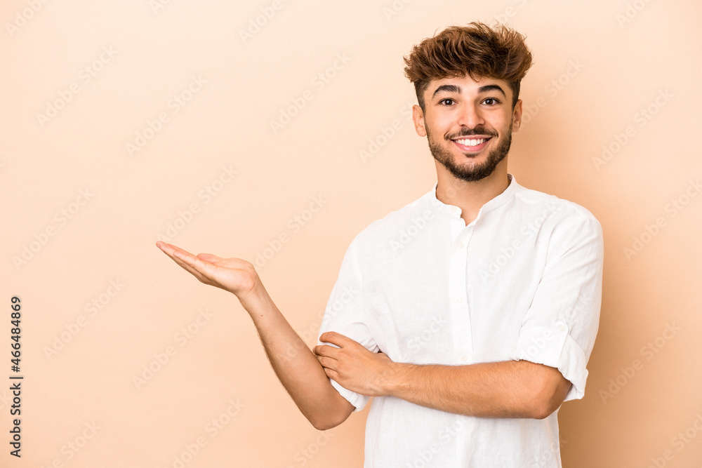 Young arab man isolated on beige background showing a copy space on a palm and holding another hand on waist. Young arab man isolated on beige background showing a copy space on a palm and holding another hand on waist.