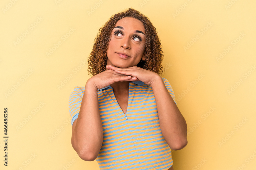 Young latin transsexual woman isolated on yellow background keeps hands under chin, is looking happily aside. Young latin transsexual woman isolated on yellow background keeps hands under chin, is looking happily aside.