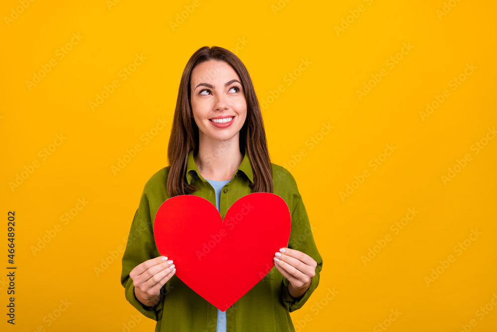 Photo of cute sweet young lady wear green shirt holding large heart looking empty space smiling isolated yellow color background Photo of cute sweet young lady wear green shirt holding large heart looking empty space smiling isolated yellow color background
