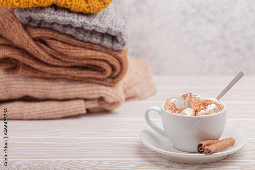 Cozy winter composition, cup with hot chocolate or cocoa with marshmallow and pile of warm knitted clothes on white wooden rustic background. Comfy and soft winter at home, free space for text Cozy winter composition, cup with hot chocolate or cocoa with marshmallow and pile of warm knitted clothes on white wooden rustic background. Comfy and soft winter at home, free space for text