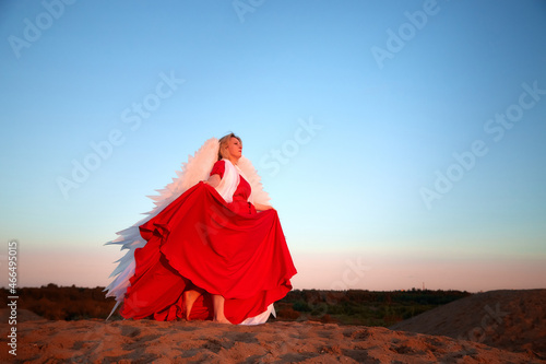 A plump blonde middle-aged woman with white angle wings in a desert with dunes and sand in a nice summer or spring sunny day with blue sky