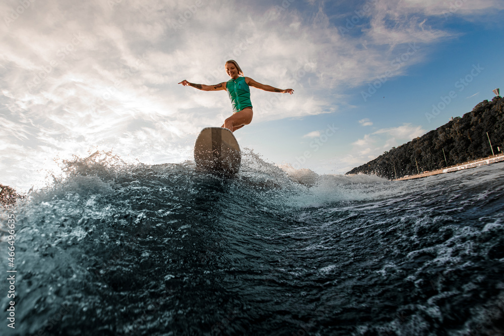 Smiling woman having fun wakesurfing on the board on the river wave against blue sky