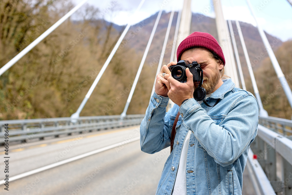 Hipster male in denim jacketwalking on bridge in countryside using camera, stand outdoors taking photo, in casual denim jacket, hat. portrait, copy space Hipster male in denim jacketwalking on bridge in countryside using camera, stand outdoors taking photo, in casual denim jacket, hat. portrait, copy space