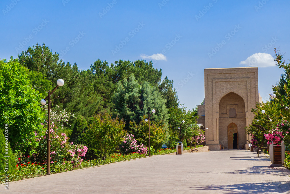 Park alley lushly planted with trees and flowering bushes, leading to main entrance of Al Hakim At-Termezi mosque (its portal is visible). Shot in Termez, Uzbekistan
