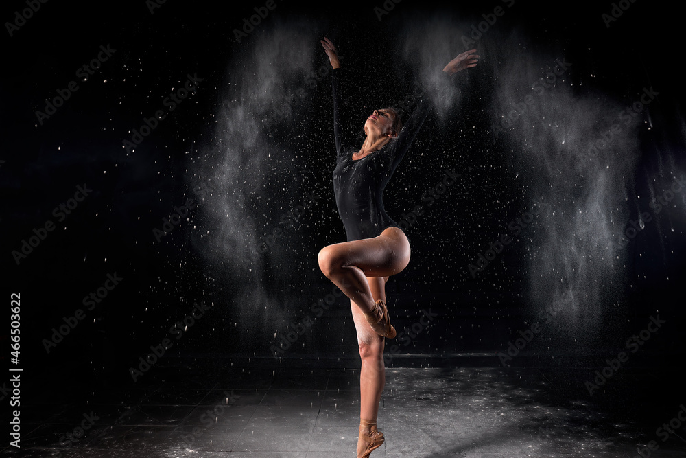Artistic dance pose using powder or dust. Beautiful expressive ballet dancer un black swimsuit in dust at studio on black background.Slim caucasian Ballerina with flour. Full-length portrait